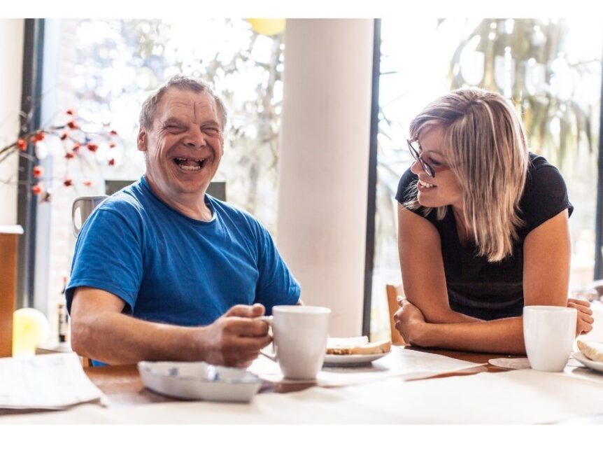 Older laughing man with Down Syndrome at table with young woman having a cup of tea.