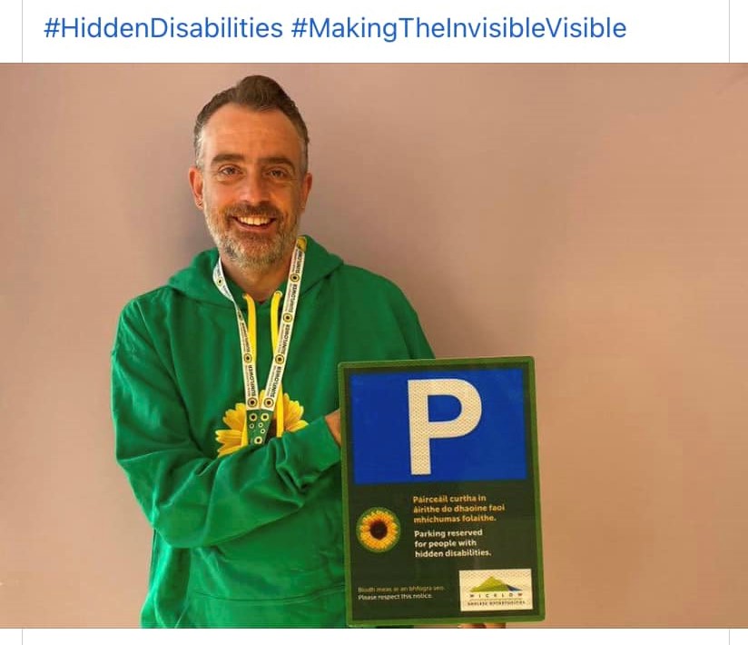 Photo of smiling man with sunflower logo beside one of the new Hidden Disabilities parking signs.