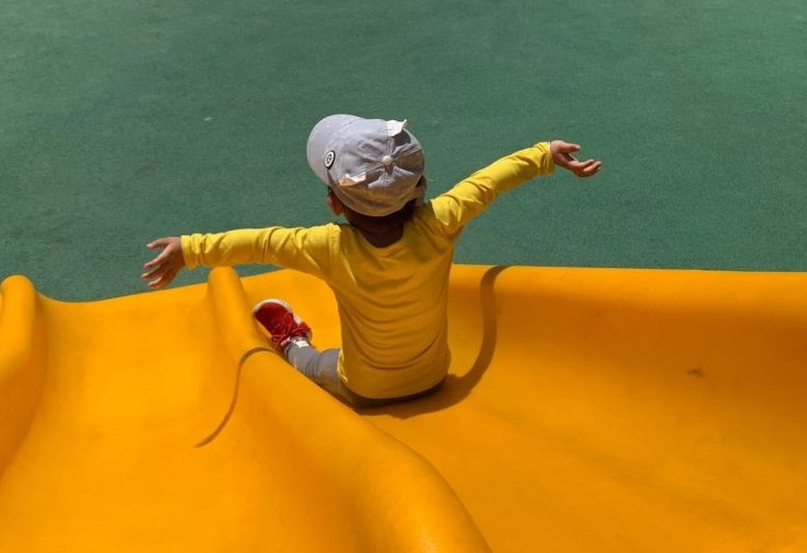 Young child going down yellow slide with arms outstretched.